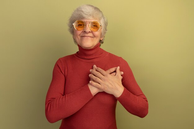 Pleased old woman wearing red turtleneck sweater and sunglasses looking at front putting hands on chest isolated on olive green wall with copy space