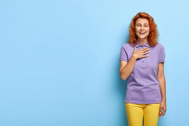Pleased overjoyed millennial woman with wavy red hair posing against the blue wall
