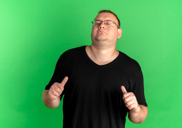 緑の壁の上に立っている自分を指している黒いTシャツを着て眼鏡をかけた太りすぎの男を喜ばせる