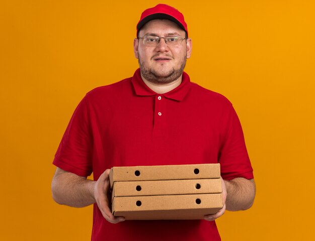 довольный толстый молодой доставщик в оптических очках держит коробки для пиццы, изолированные на оранжевой стене с копией пространства