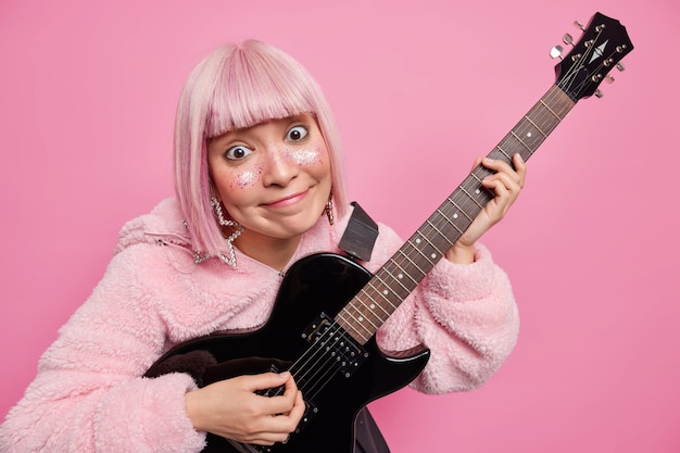 Pleased pink haired woman plays electric guitar performs favorite genre of music has face decorated with glitters dressed in coat 