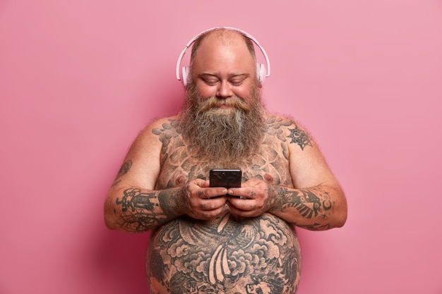 Довольный пухлый мужчина с обнаженным татуированным телом, большим животом, слушает музыку в наушниках, держит сотовый, скачивает песни в плейлист, изолирован на розовой стене. Люди, избыточный вес, концепция хобби
