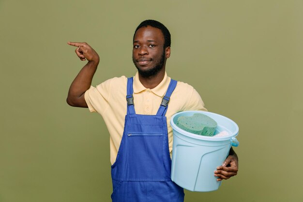 Pleased points at side holding bucket young africanamerican cleaner male in uniform with gloves isolated on green background