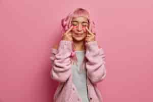 Free photo pleased positive woman has long rosy hair, makes curly hairstyle with curlers, indicates at beauty patches, stands with closed eyes wears warm sweater