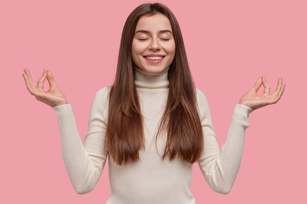 Pleased positive young European woman makes mudra gesture, keeps eyes shut, relaxes after work, dressed in casual clothes, isolated over pink background. People, wellness