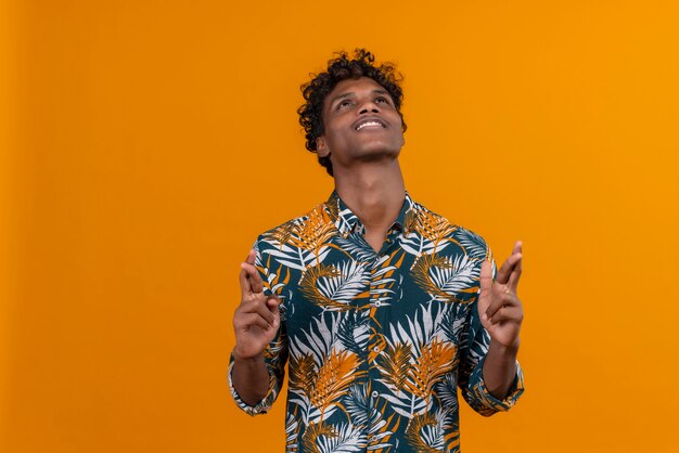 Довольный и позитивный молодой красивый темнокожий мужчина с вьющимися волосами в рубашке с принтом листьев, держащей пальцы вместе, глядя вверх на оранжевом фоне