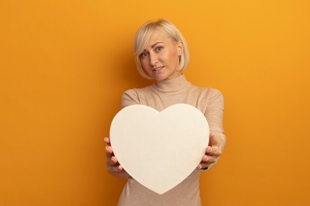 Pleased pretty blonde slavic woman holds heart shape on orange