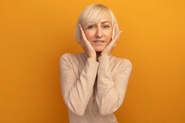 Pleased pretty blonde slavic woman puts hands on face isolated on orange wall