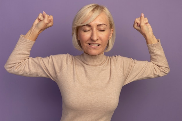 Free photo pleased pretty blonde slavic woman stands with closed eyes raising fists up on purple