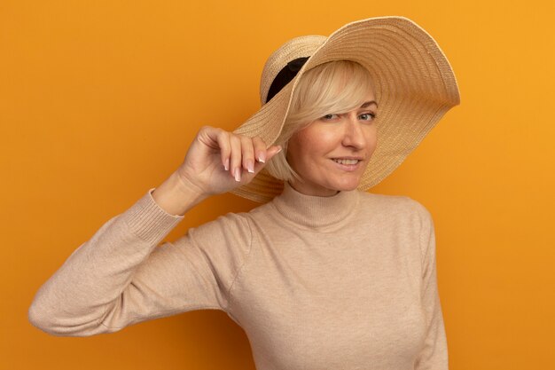 Pleased pretty blonde slavic woman with beach hat holds hat on orange