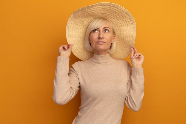 Pleased pretty blonde slavic woman with beach hat looks up on orange