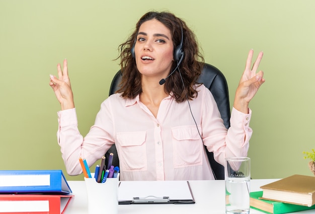 Piacevole operatore di call center femminile caucasica sulle cuffie seduto alla scrivania con strumenti da ufficio che gesticolano segno di vittoria