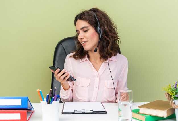 Довольная женщина-оператор колл-центра в наушниках сидит за столом с офисными инструментами и смотрит на телефон, изолированный на зеленой стене