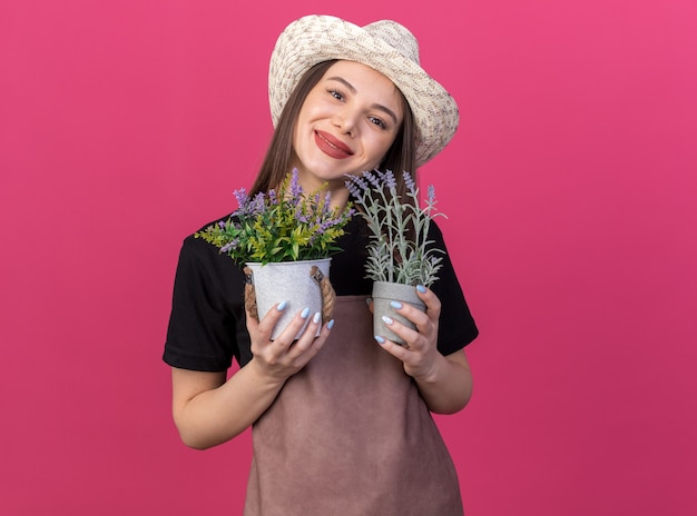 Pleased pretty caucasian female gardener wearing gardening hat holding flowerpots isolated on pink wall with copy space