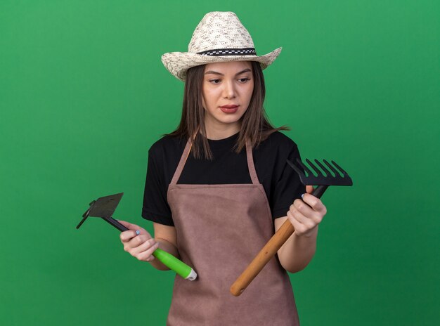 Pleased pretty caucasian female gardener wearing gardening hat holding hoe rake and looking at rake