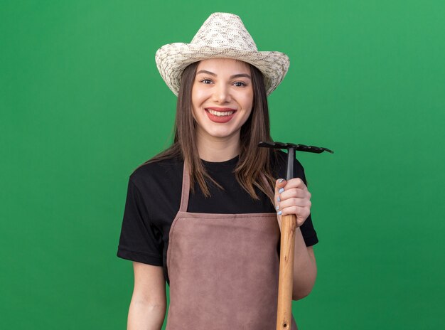 Pleased pretty caucasian female gardener wearing gardening hat holding rake on green