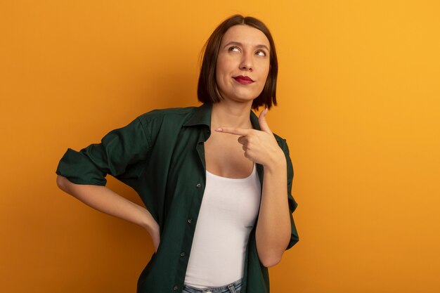 Pleased pretty caucasian woman points at side looking up on orange