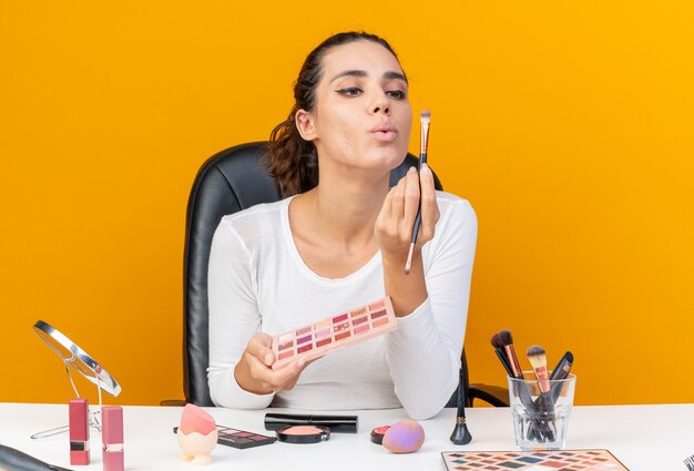 Довольная красивая кавказская женщина сидит за столом с инструментами для макияжа, держит палитру теней и смотрит на кисти для макияжа, изолированные на оранжевой стене с копией пространства
