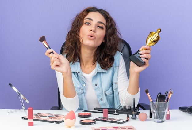 Pleased pretty caucasian woman sitting at table with makeup tools holding makeup brush and winner cup