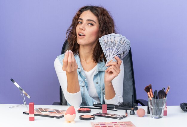 Pleased pretty caucasian woman sitting at table with makeup tools holds money isolated on purple wall with copy space