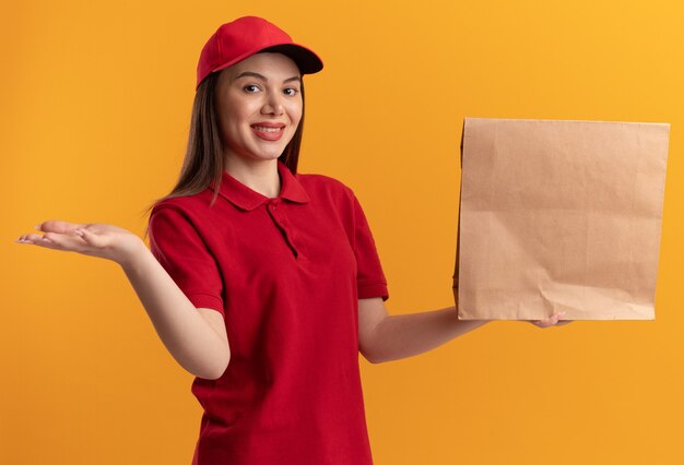 Pleased pretty delivery woman in uniform keeps hand open and holds paper package on orange