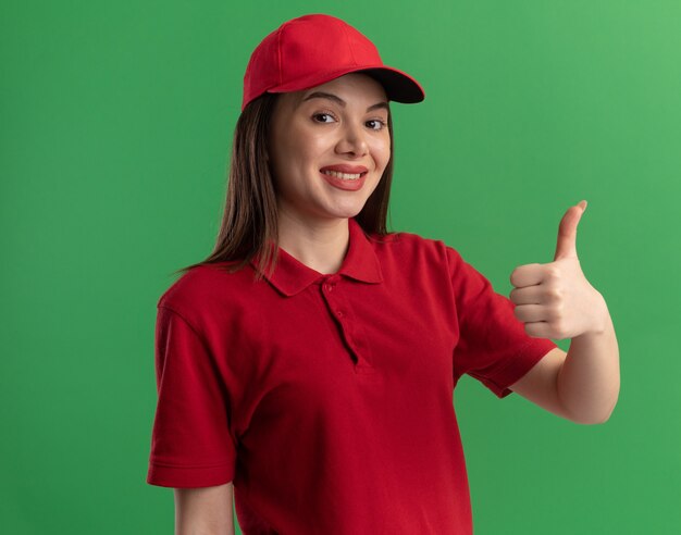 Pleased pretty delivery woman in uniform thumbs up on green