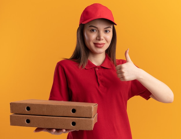 Pleased pretty delivery woman in uniform thumbs up and holds pizza boxes isolated on orange wall with copy space