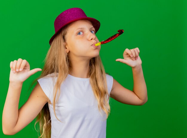 Pleased pretty little girl in holiday hat blowing whistle celebrating birthday