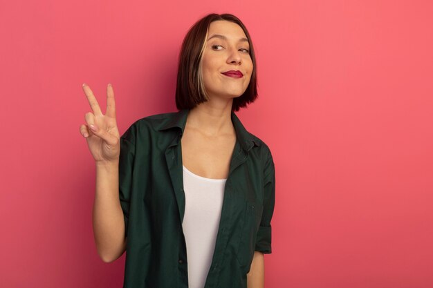 Pleased pretty woman gestures victory hand sign looking at side isolated on pink wall