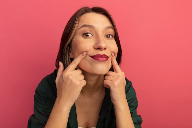 Pleased pretty woman keeps smile with fingers isolated on pink wall