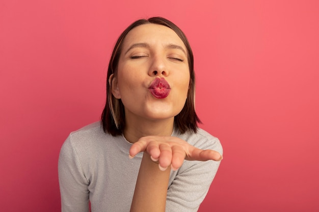 La donna graziosa soddisfatta invia il bacio con la mano isolata sulla parete rosa