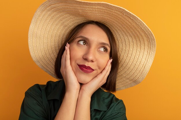 Pleased pretty woman with beach hat puts hands on face isolated on orange wall