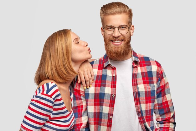 Pleased pretty woman with bobbed hairstyle, stands sideways, leans at shoulder of her handsome bearded boyfriend