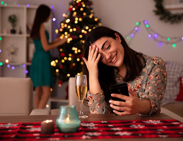 기쁘게 꽤 어린 소녀가 크리스마스 트리를 장식하고 그녀의 친구가 이마에 손을 대고 테이블에 앉아 집에서 크리스마스 시간을 즐기는 전화를 봅니다.