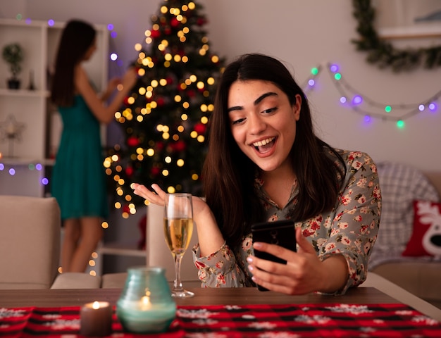 pleased pretty young girl decorates christmas tree and her surprised friend looks at phone sitting at table and enjoying christmas time at home