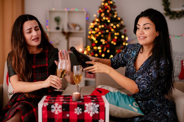 肘掛け椅子に座って家でクリスマスの時間を楽しんでいる彼女のグラスにシャンパンを注ぐ友人に手で十分に満足しているかなり若い女の子のジェスチャー