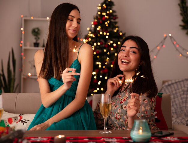 기쁘게 예쁜 젊은 여자가 폭죽을 보유하고 테이블에 앉아 집에서 크리스마스 시간을 즐기는 그녀의 친구를 본다