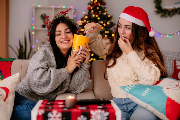 Pleased pretty young girl with holly wreath holds popcorn bucket and her friend with santa hat eat popcorn sitting on armchair and enjoying christmas time at home