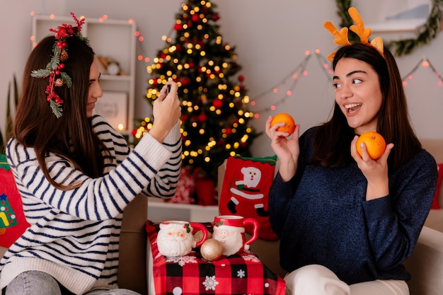 Pleased pretty young girl with holly wreath takes photo of her friend holding oranges sitting on armchair and enjoying christmas time at home