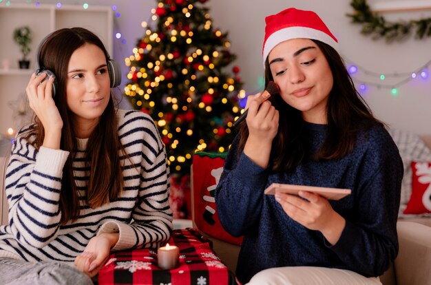 Довольная красивая молодая девушка в шляпе санта-клауса делает макияж, сидя на кресле со своим другом в наушниках, наслаждаясь Рождеством дома
