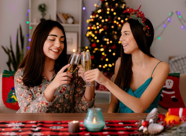 pleased pretty young girls clink glasses of champagne sitting at table and enjoying christmas time at home