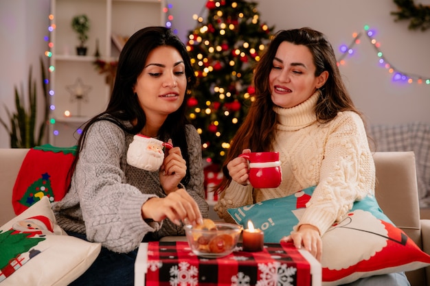 Le ragazze graziose e contente tengono le tazze e guardano i biscotti seduti sulle poltrone e si godono il periodo natalizio a casa