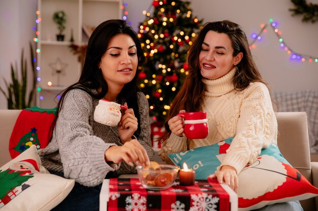 Pleased pretty young girls hold cups and look at biscuits sitting on armchairs and enjoying christmas time at home