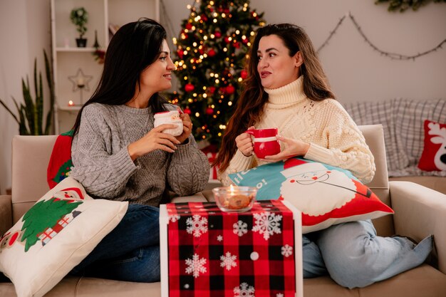 pleased pretty young girls hold cups and look at each other sitting on armchairs and enjoying christmas time at home