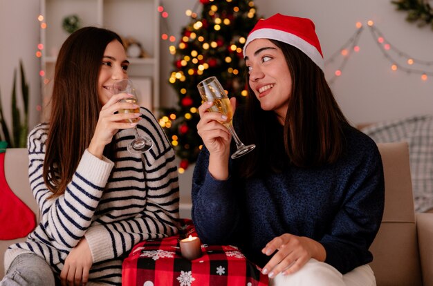 pleased pretty young girls hold glasses of champagne and look at each other sitting on armchairs and enjoying christmas time at home