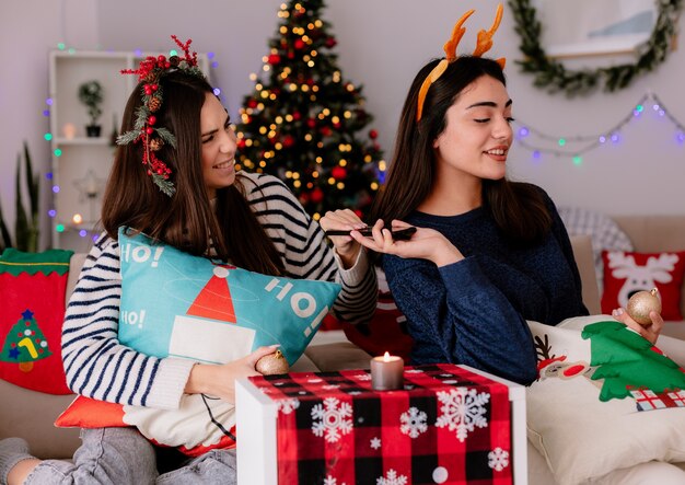 ヒイラギの花輪とトナカイのヘッドバンドを持ったかなり若い女の子が、肘掛け椅子に座って家でクリスマスの時間を楽しんでいるガラス玉の飾りと電話を持って喜んでいます