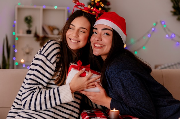순록 안경과 산타 모자와 함께 기쁘게 예쁜 어린 소녀는 안락 의자에 앉아 집에서 크리스마스 시간을 즐기는 크리스마스 선물 상자를 개최