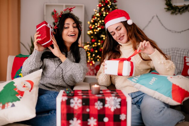 Pleased pretty young girls with santa hat and holly wreath hold gift boxes sitting on armchairs and enjoying christmas time at home
