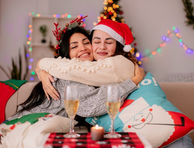 산타 모자와 함께 기쁘게 예쁜 어린 소녀는 안락 의자에 앉아 집에서 크리스마스 시간을 즐기고 서로 포옹