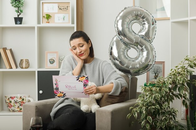 Pleased putting hand on cheek beautiful girl on happy women day holding and reading greeting card sitting on armchair in living room